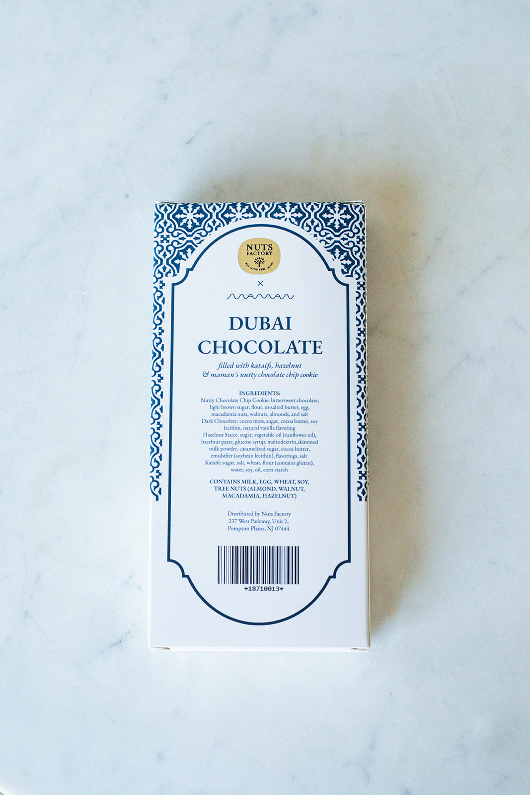 A rectangular box of Nut Factory Dubai Chocolate, featuring a white background with a blue ornate border and black text, photographed flat on a light marble surface.