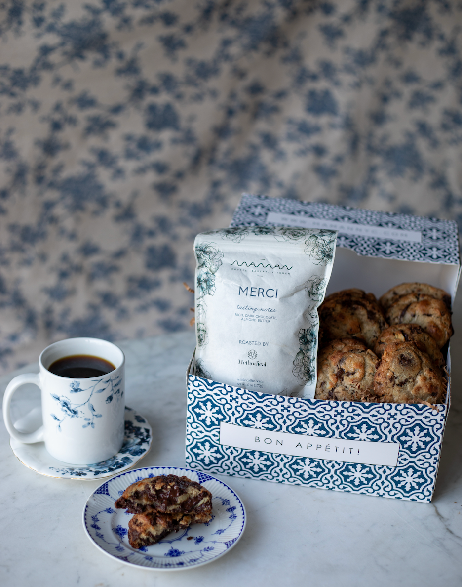 blue and white pastry gift box with assorted nutty chocolate chip cookies and Maman Merci coffee, shown on a table with a cup of coffee and a plate with a cookie.