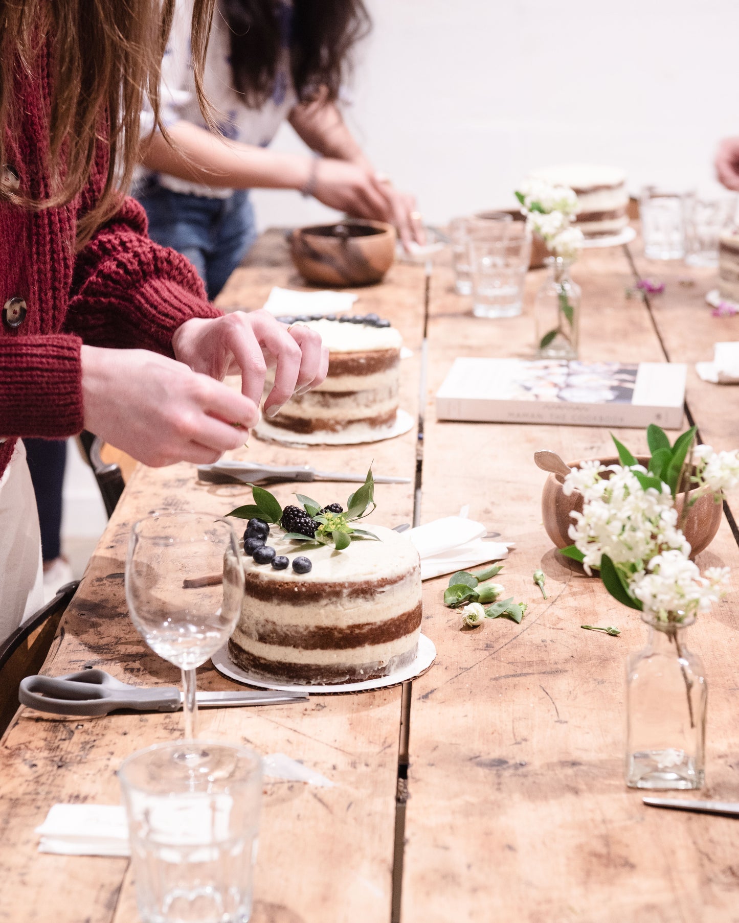 People decorating a layered cake with blueberries and flowers.