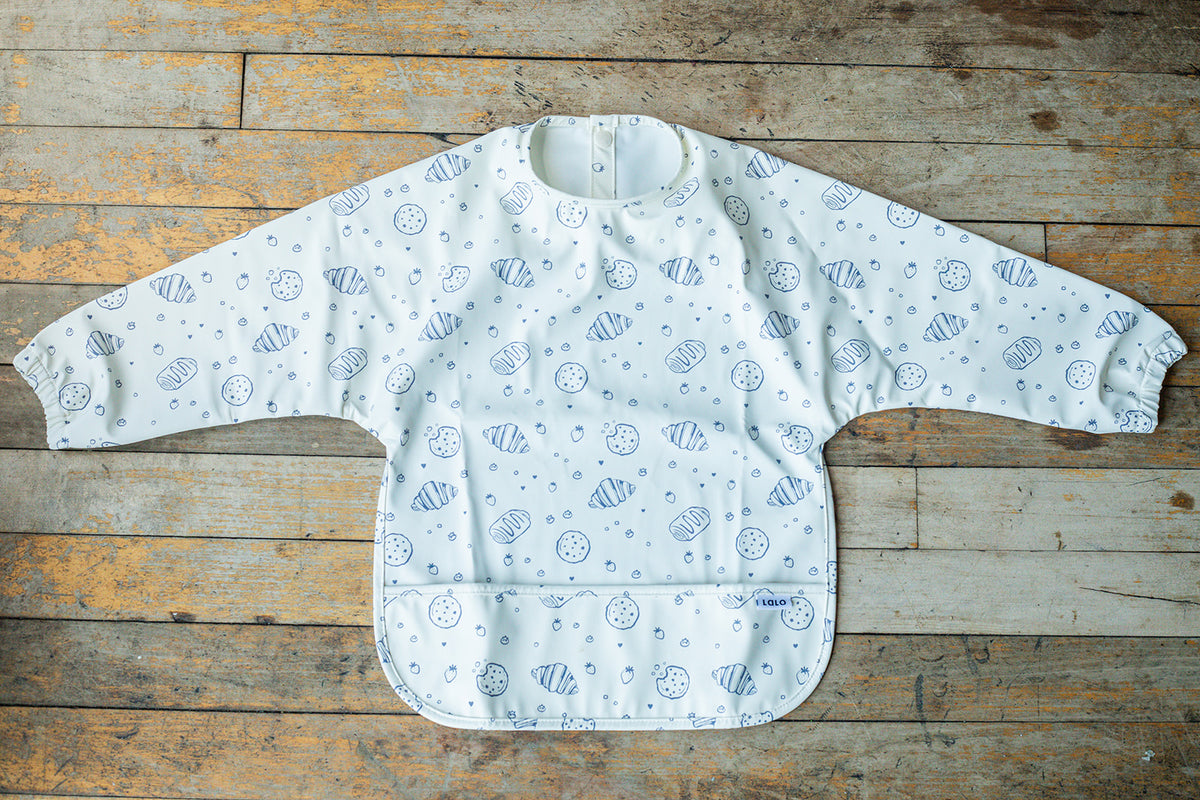 A long-sleeve baby bib with a white background and a blue seashell and dot pattern, laid flat on a rustic wooden plank surface.