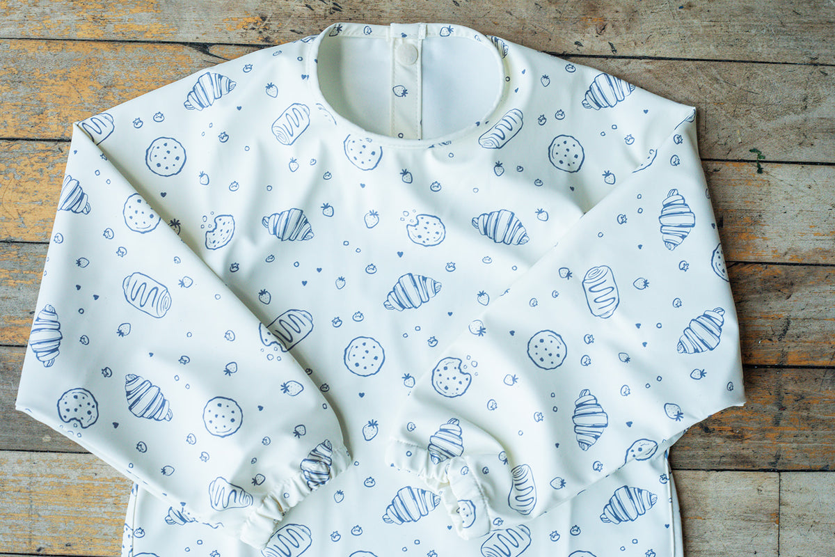 A white long-sleeve baby bib with a blue sketch-style print of croissants, pastries, and dots, laid flat on a rustic wooden surface.