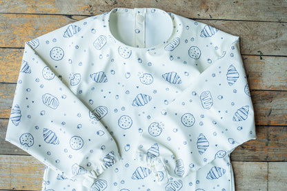 A white long-sleeve baby bib with a blue sketch-style print of croissants, pastries, and dots, laid flat on a rustic wooden surface.