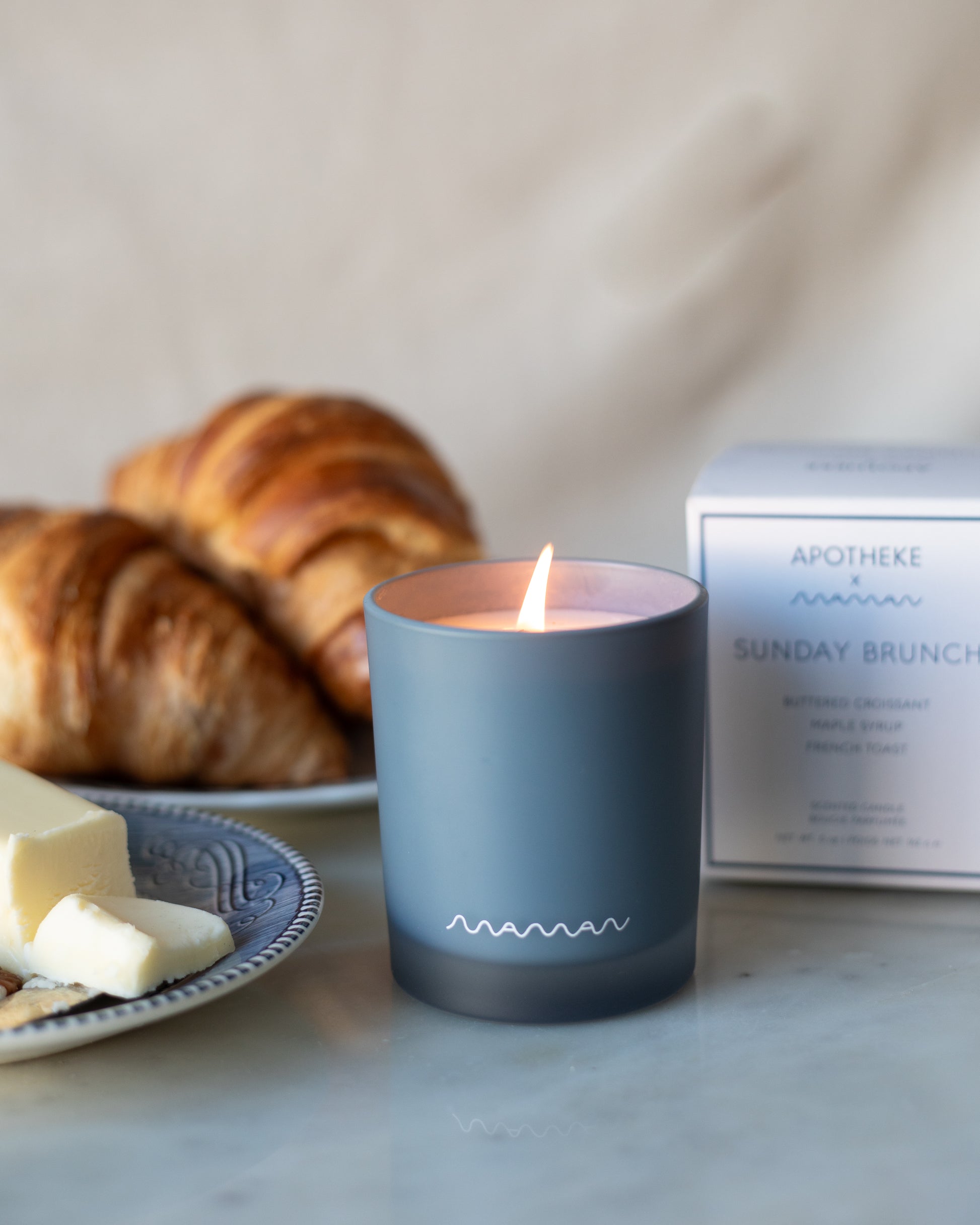Blue Apotheke candle with white maman logo, in front of white box labeled 'Sunday Brunch', with croissants and butter. 