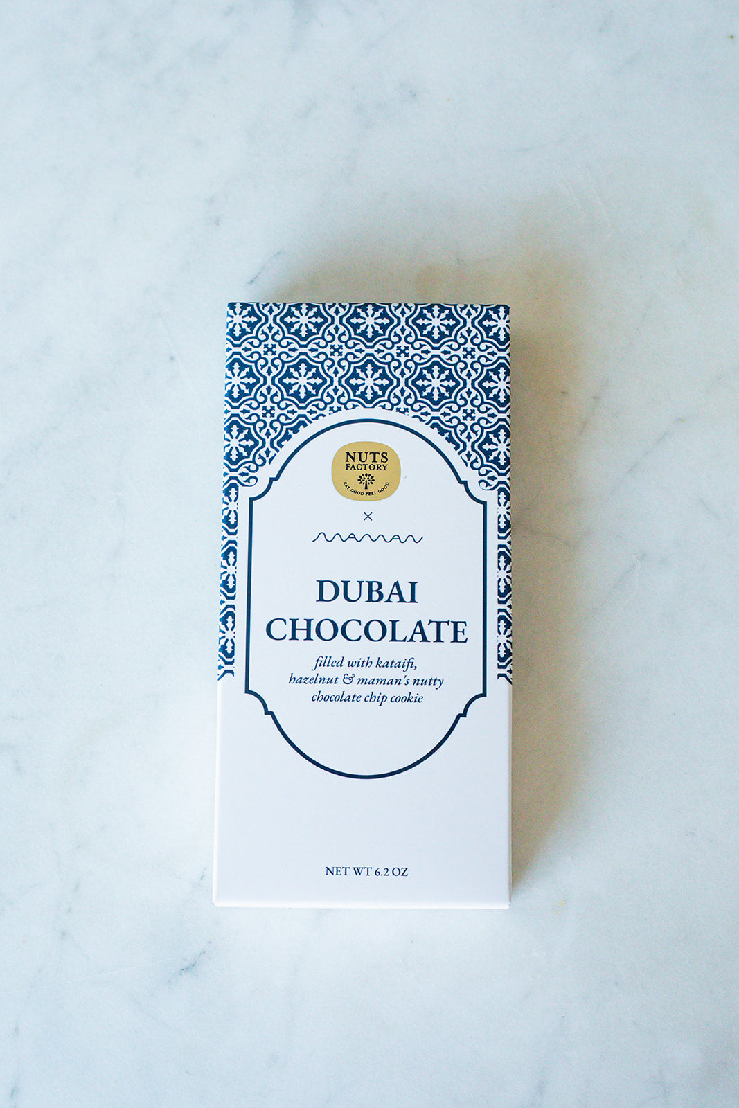 A boxed bar of Nuts Factory Dubai Chocolate, featuring a white box with a blue geometric pattern border and a gold logo seal, set on a light marble surface.
