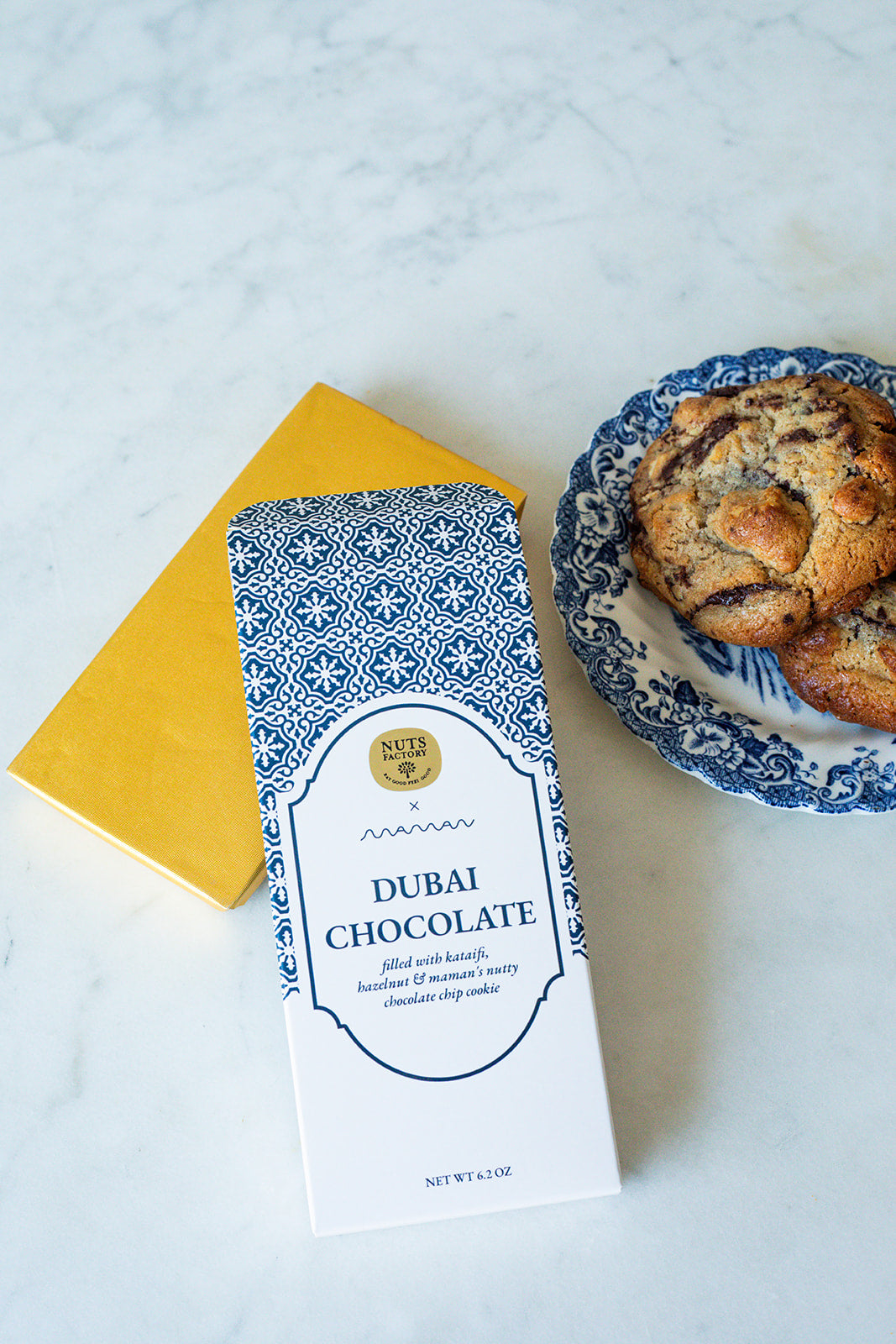 A package of Nuts Factory Dubai Chocolate cookies lies on a white marble surface next to a yellow notebook and a blue-and-white patterned plate with chocolate chip cookies.