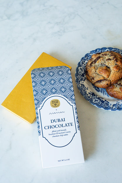 A package of Nuts Factory Dubai Chocolate cookies lies on a white marble surface next to a yellow notebook and a blue-and-white patterned plate with chocolate chip cookies.