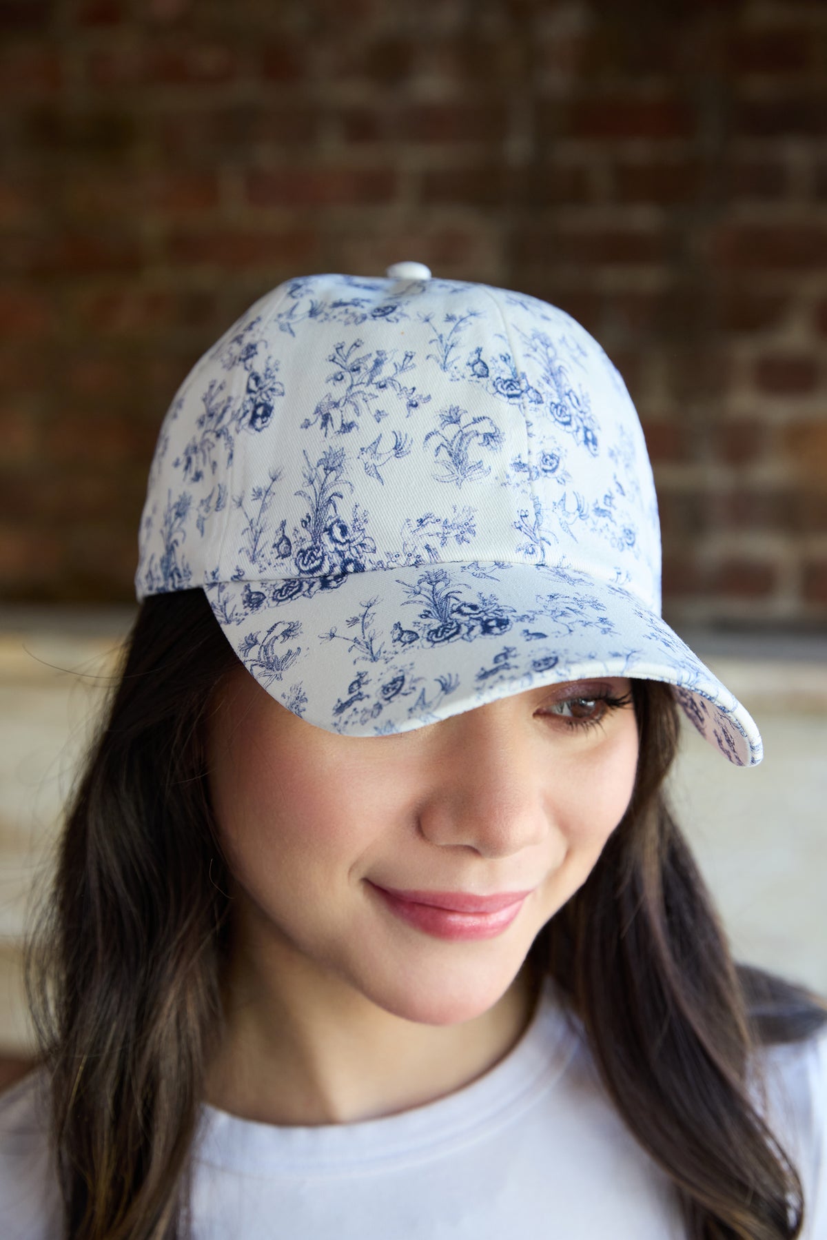 A woman wearing a white baseball cap with a blue floral toile pattern, photographed in a lifestyle setting with a blurred brick wall background.