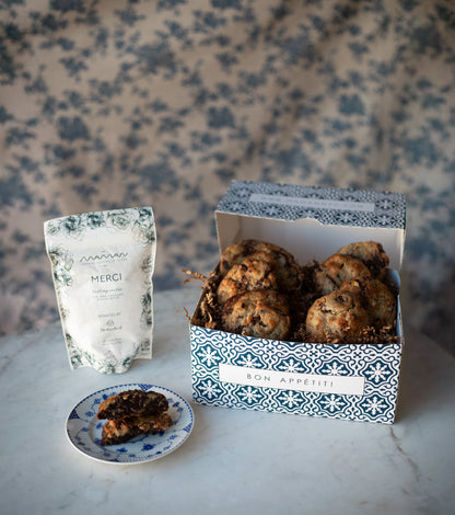 blue and white pastry gift box with assorted nutty chocolate chip cookies and Maman Merci coffee, shown on a table with a plate with a cookie.