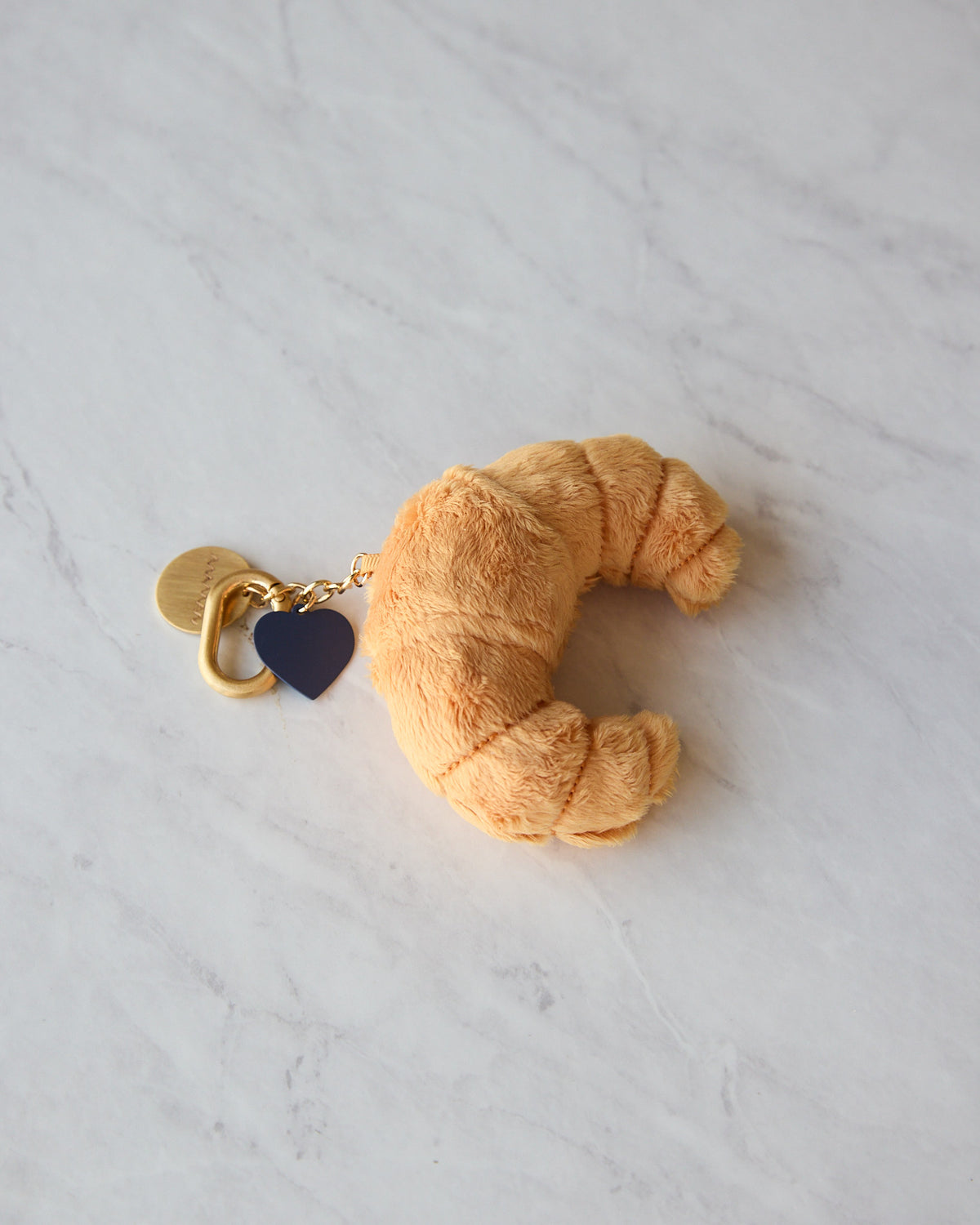 A plush croissant-shaped keychain or bag charm in a warm tan color, attached to a gold-tone ring with a small dark navy heart charm, resting on a white marble surface.