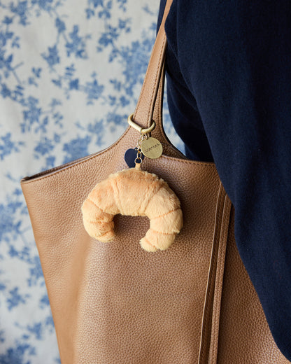 A close-up of a tan pebbled leather tote bag with a croissant-shaped plush keychain charm hanging from the strap. The bag is being carried by someone wearing a navy blue top, with a blue and white floral patterned fabric in the background.