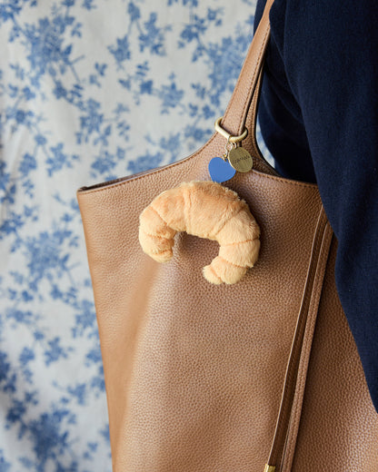 A tan pebbled leather tote bag with a croissant-shaped plush keychain charm and small blue and gold heart charms, shown being carried on a person's shoulder against a blue and white floral background.