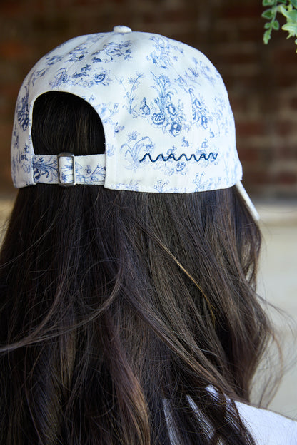 A close-up back view of a person wearing a white baseball cap with a blue floral pattern and blue embroidered script on the side, with long dark brown hair visible.
