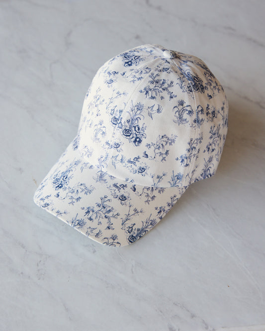 A baseball cap with a blue floral toile pattern on a white fabric, displayed on a light gray marble surface.