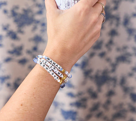 Three bracelets worn on the wrist, each with a gold croissant bead and letter beads spelling a different word “bonjour”, “maman”, and “je t’aime”, in shades of blue, blue-and-white, and white-and-gold beads.