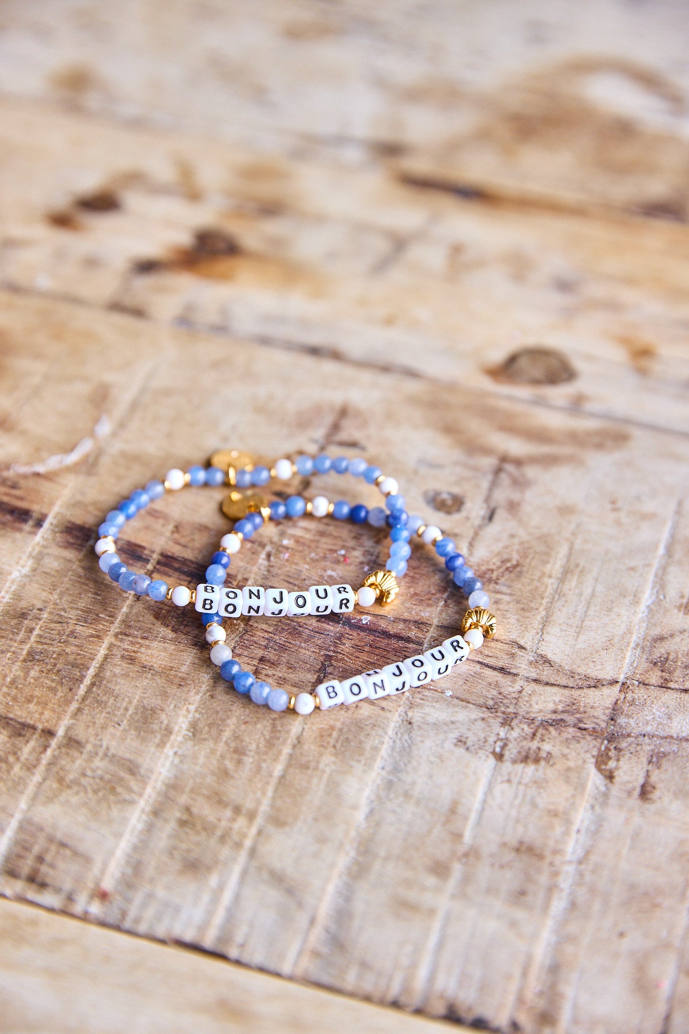 Bracelet with shades of blue beads, a gold croissant bead, and letter beads spelling “bonjour”.