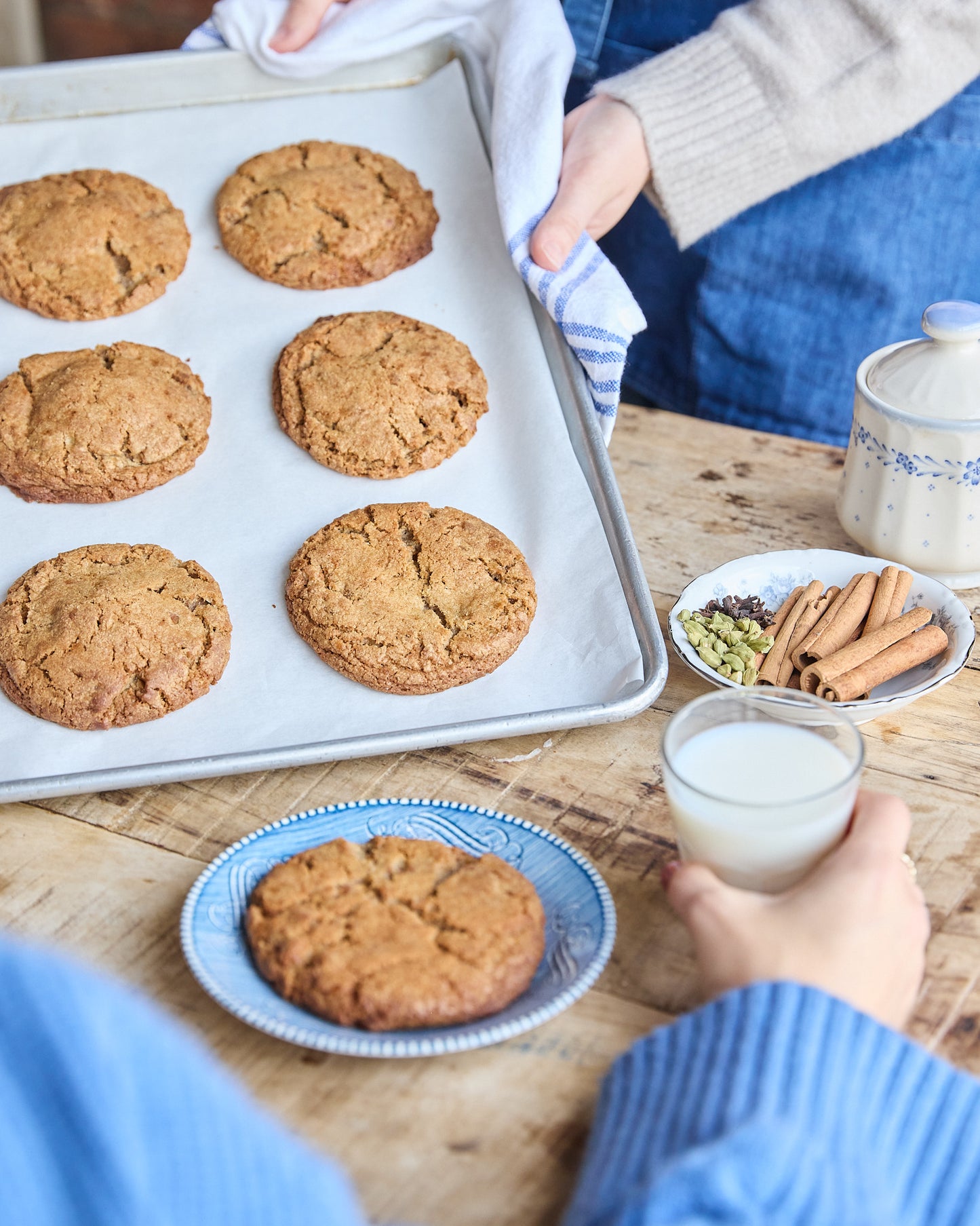 Baked cookies on a tray with a person holding a glass of milk, surrounded by spices and a plate of cookies.