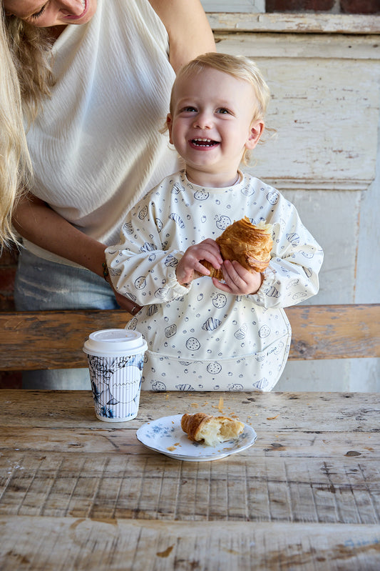 A toddler wearing a cream long-sleeve bib with a light blue smiley face pattern sits at a rustic wooden table, holding a croissant. A coffee cup and a plate with a croissant are on the table, and a woman is partially visible behind the child.