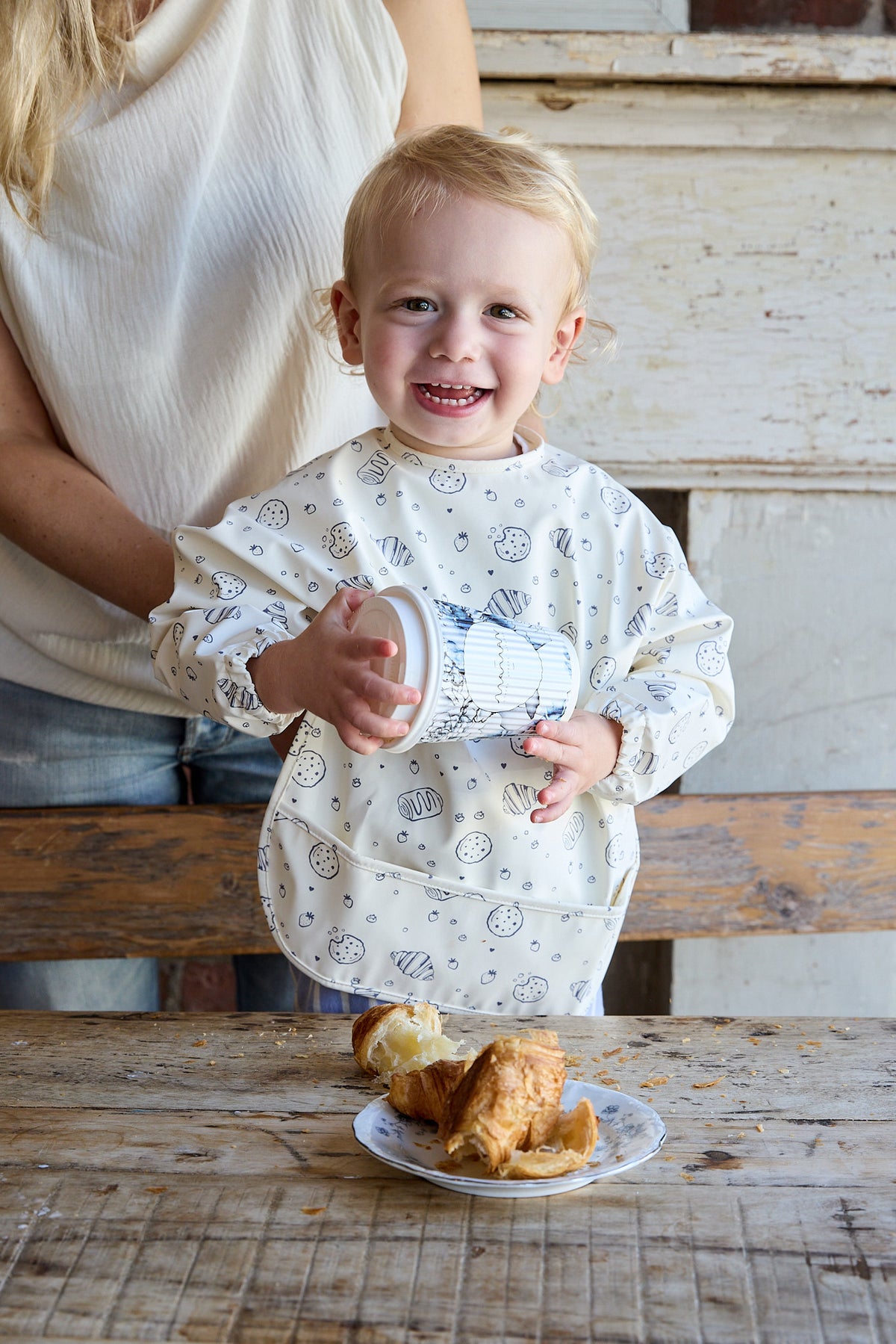 A smiling young child wearing a long-sleeve bib stands at a rustic wooden table holding a small cup, with a plate of pastries in front and part of an adult visible behind them.