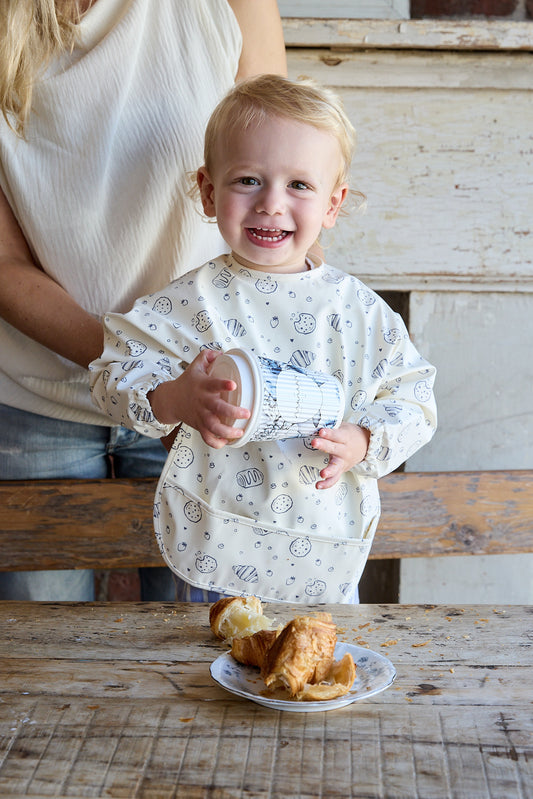 A smiling young child wearing a long-sleeve bib stands at a rustic wooden table holding a small cup, with a plate of pastries in front and part of an adult visible behind them.