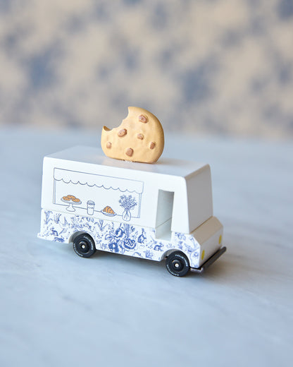 A miniature white toy food truck with blue floral patterns and black wheels, topped with a small broken cookie, set against a soft-focus neutral background.