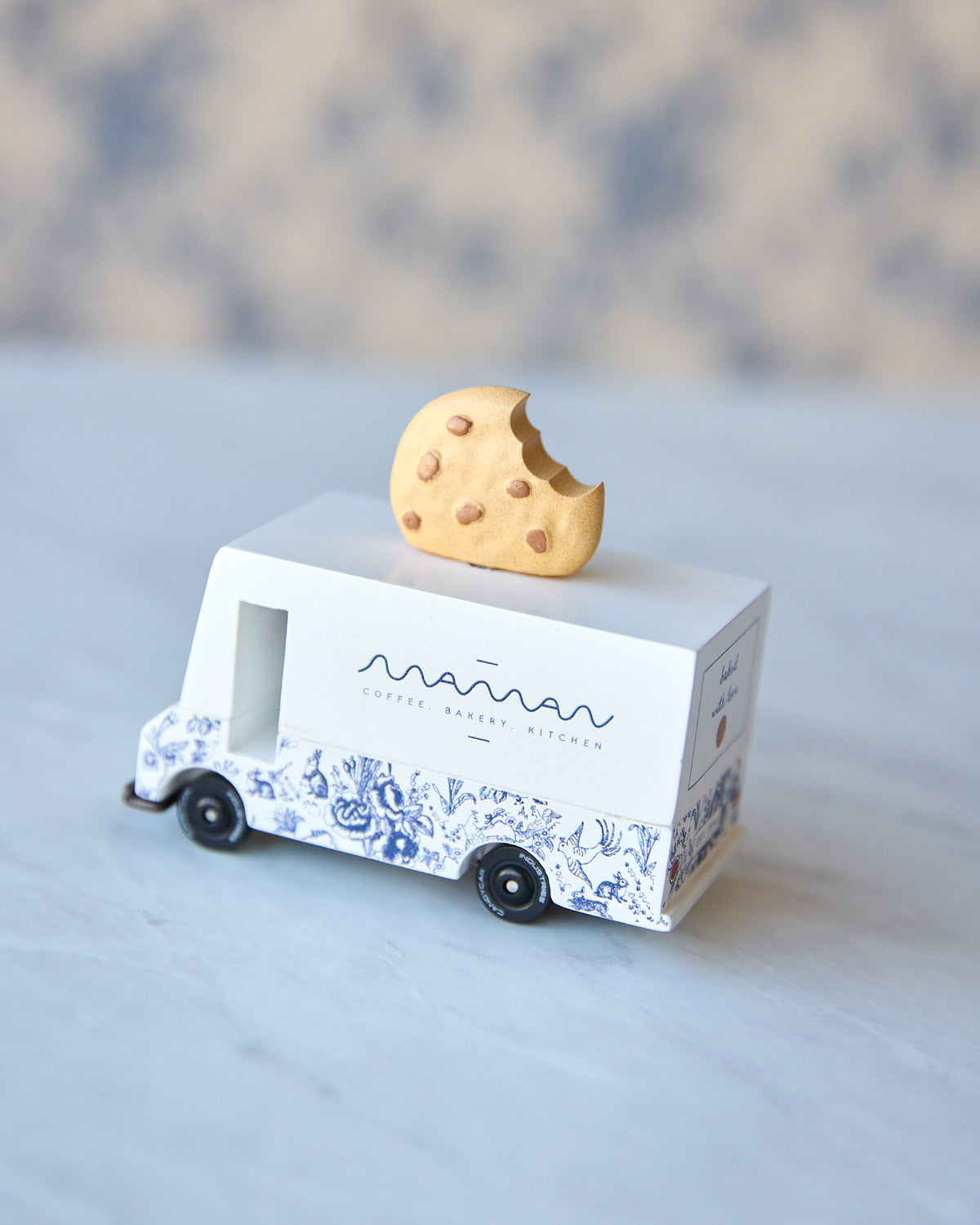 A small white toy delivery truck with blue floral patterns and the text 'mama cookie bakery' on the side, with a half-eaten chocolate chip cookie balanced on its roof, set against a soft-focus neutral background.