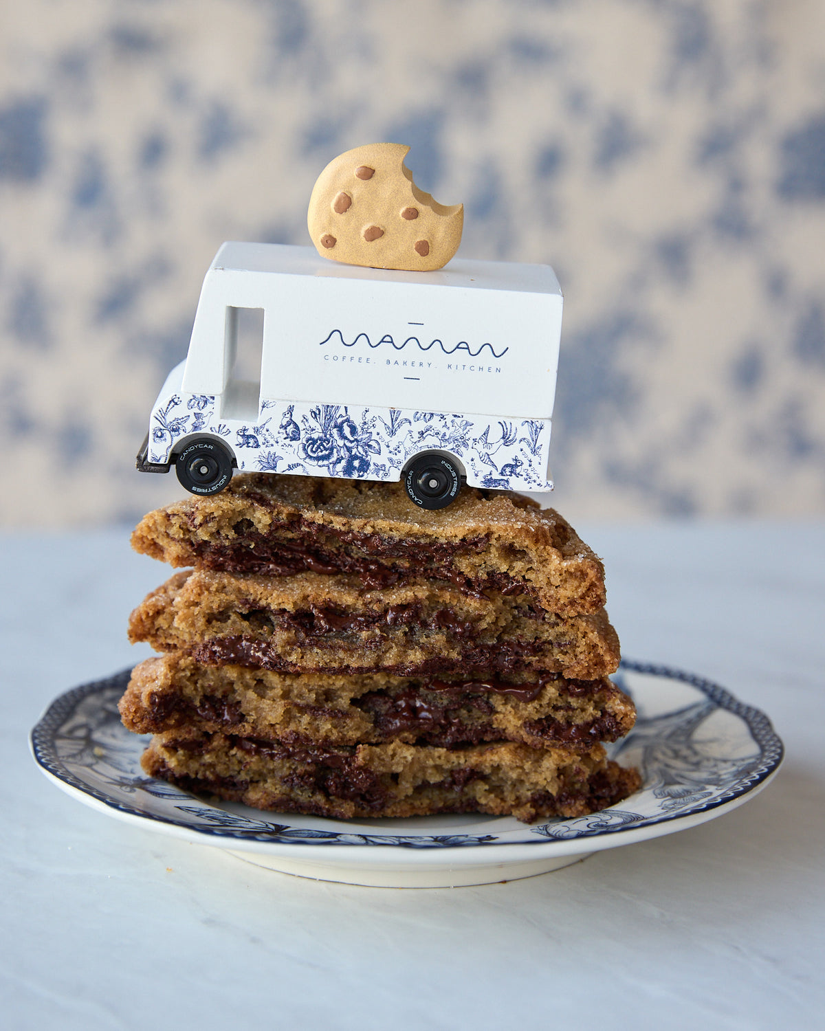 A stack of chocolate chip cookies sits on a blue-and-white patterned plate, with a small white delivery truck toy and a cookie on top. The truck is branded 'maman' with 'CHOCOLAT CHIP COOKIES' written below.