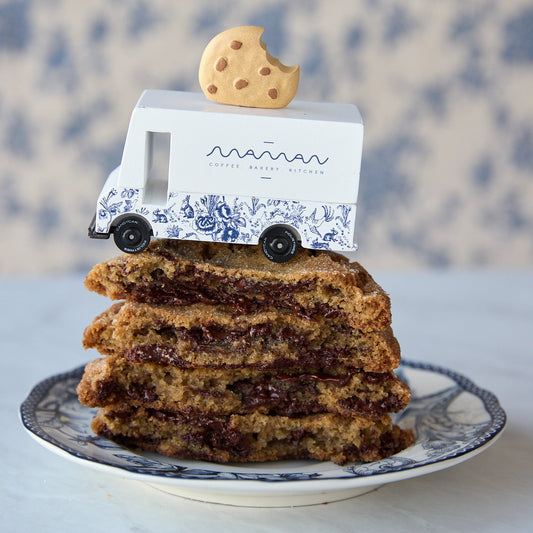 A stack of chocolate chip cookies sits on a blue-and-white patterned plate, with a small white delivery truck toy and a cookie on top. The truck is branded 'maman' with 'CHOCOLAT CHIP COOKIES' written below.