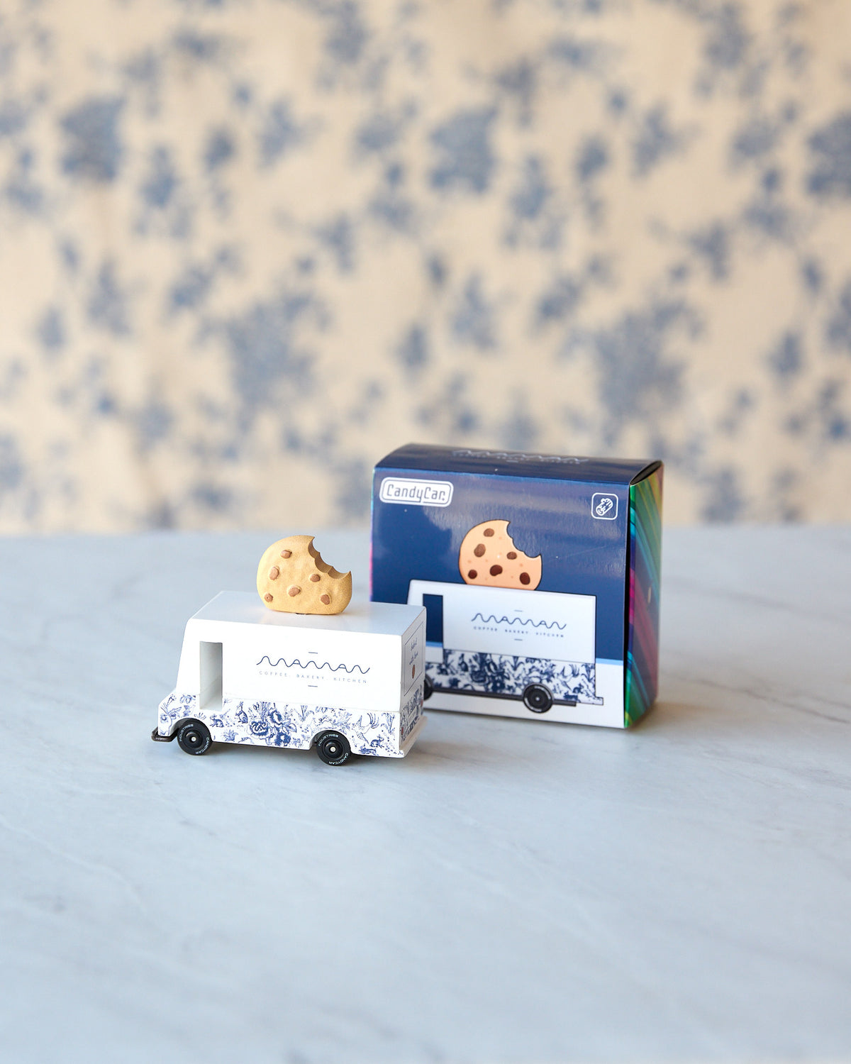 A small white toy food truck with blue illustrations and a cookie on top, shown next to its blue product box on a light marble surface with a blue floral background.