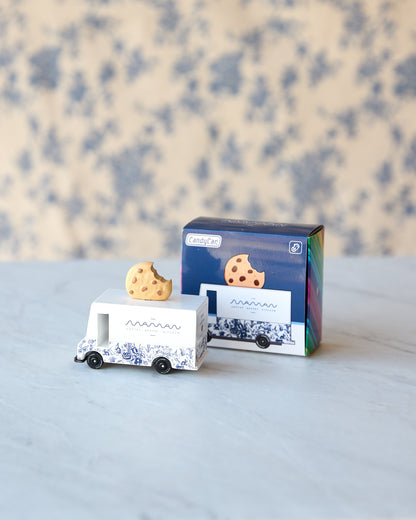 A small white toy food truck with blue illustrations and a cookie on top, shown next to its blue product box on a light marble surface with a blue floral background.