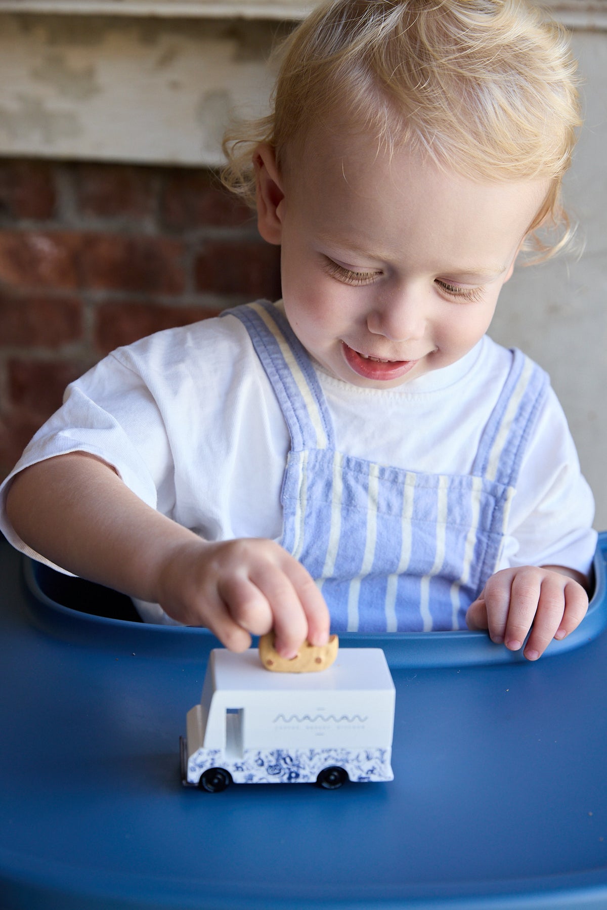 A young toddler with blonde hair wearing a white shirt and blue-and-white striped overalls sits in a blue high chair, playing with a small white toy truck that has blue floral patterns and a yellow roof.