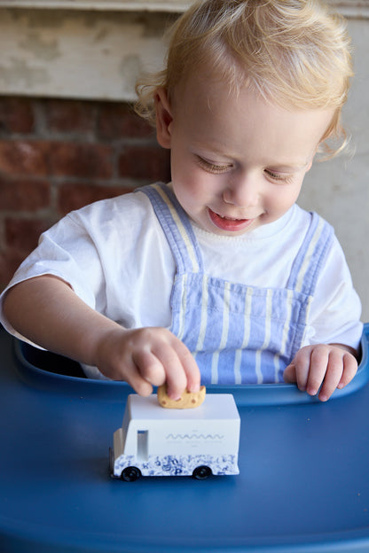 A young toddler with blonde hair wearing a white shirt and blue-and-white striped overalls sits in a blue high chair, playing with a small white toy truck that has blue floral patterns and a yellow roof.