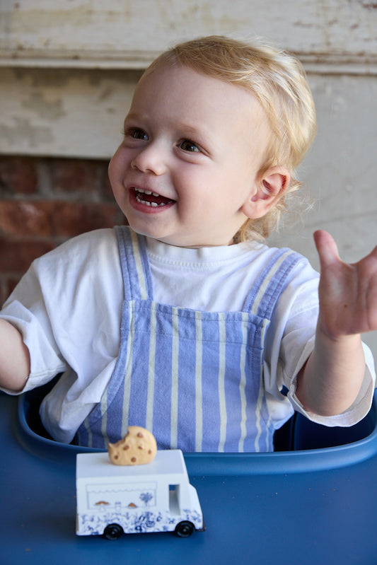 A smiling toddler with blonde hair sits in a blue high chair, wearing a white shirt and blue-and-white striped overalls. A small toy food truck with a cookie on top is in front of the child.
