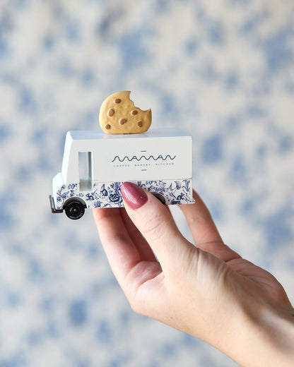 A hand with pink nail polish holds a miniature white and blue patterned food truck toy with a small cookie on its roof against a soft, blurred blue and white background.