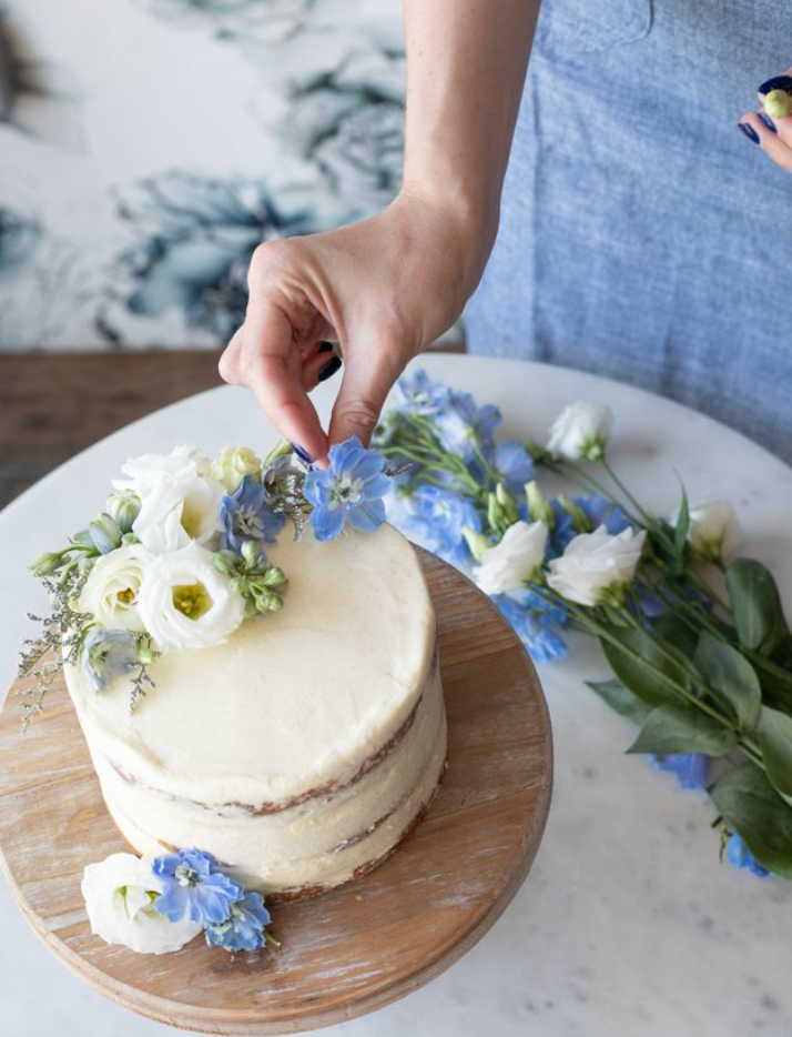 Hand placing blue flowers on a small cake with white icing. 