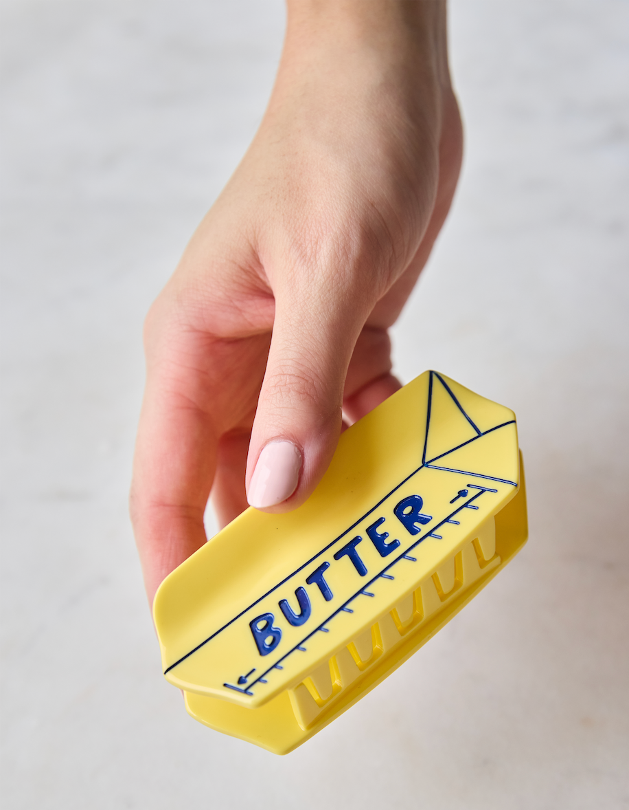 A hand holding a yellow butter dish with blue line drawings and the word 'BUTTER' printed on it, set against a light marble background.