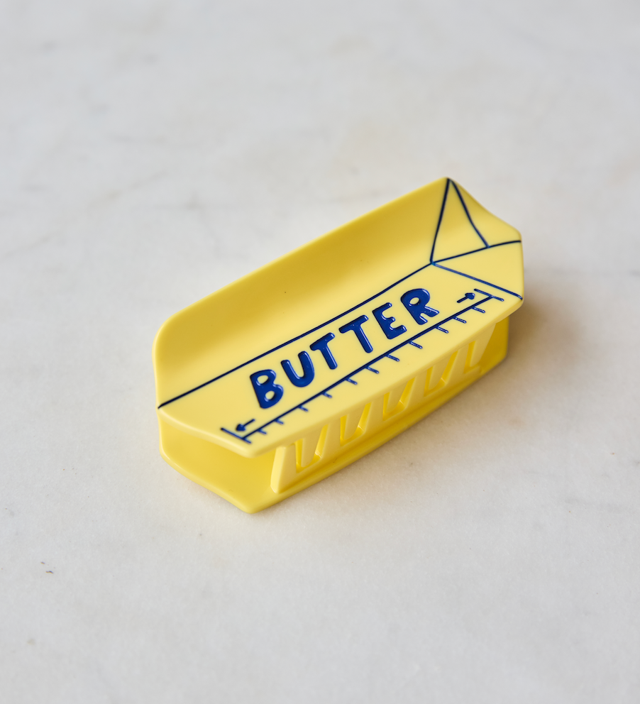 A small yellow butter dish with a printed blue ruler design and the word 'BUTTER' on the front, photographed on a light marble-like surface.