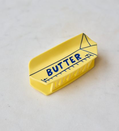 A small yellow butter dish with a printed blue ruler design and the word 'BUTTER' on the front, photographed on a light marble-like surface.